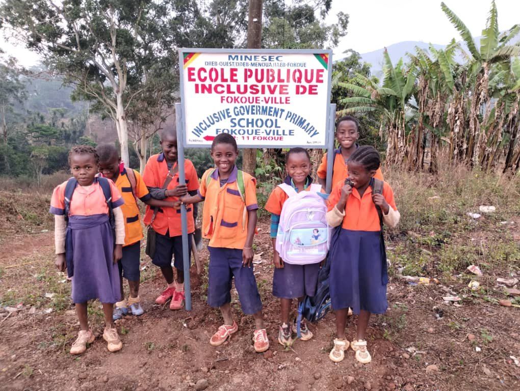 Pupils with their kits in front of the Fokoué-Ville Inclusive Public School sign